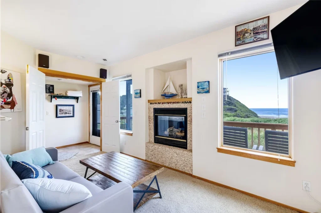 Living room with a light gray couch, wooden coffee table, stone gas fireplace, and a large window looking out to the ocean coast