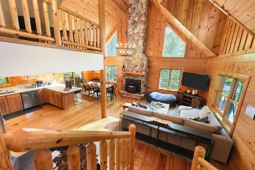 Log cabin living room with a tall stone fireplace, a loft area, and wood paneled walls