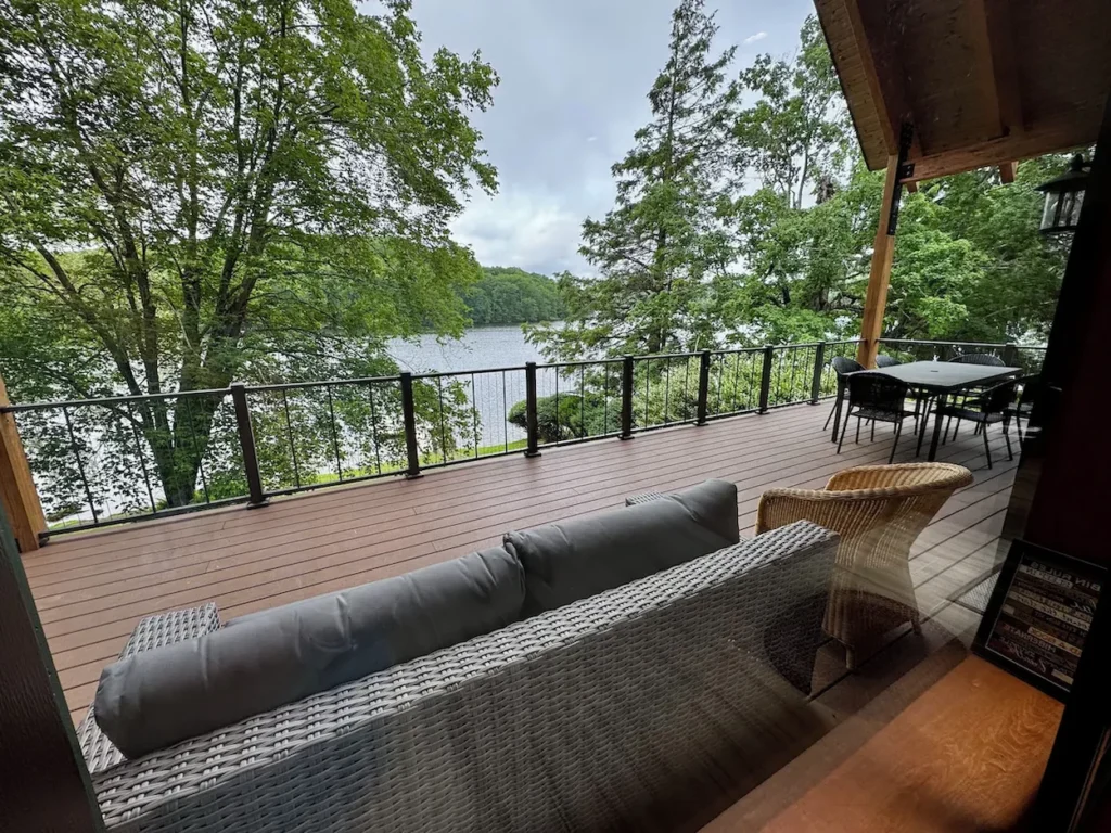 Spacious wooden cabin deck with wicker furniture and a dining table overlooking a quiet lake