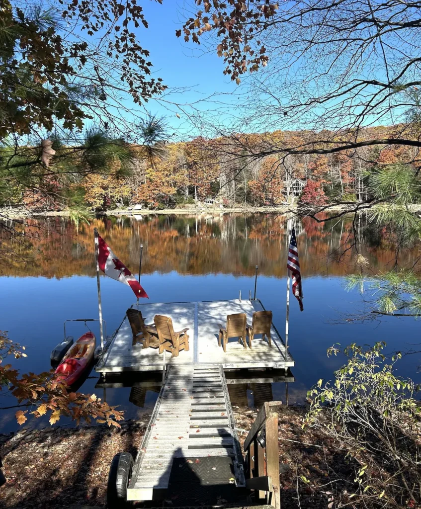 Wooden lake dock with Adirondack chairs, a Canadian flag, and an American flag surrounded by fall foliage
