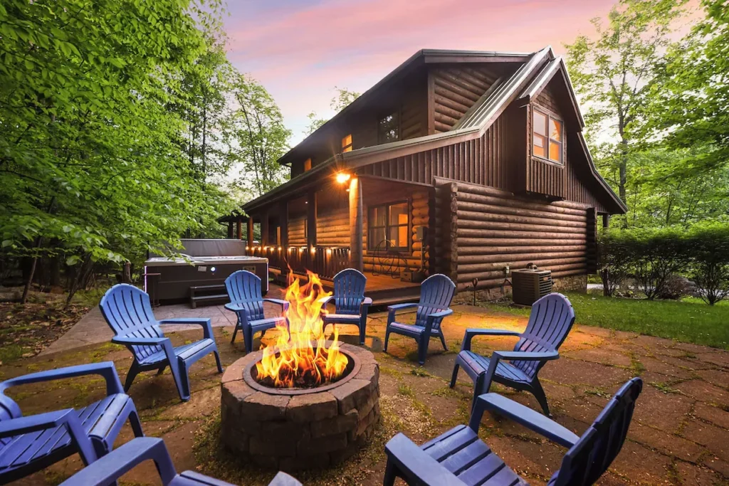 Stone fire pit with blue Adirondack chairs outside a log cabin with a hot tub