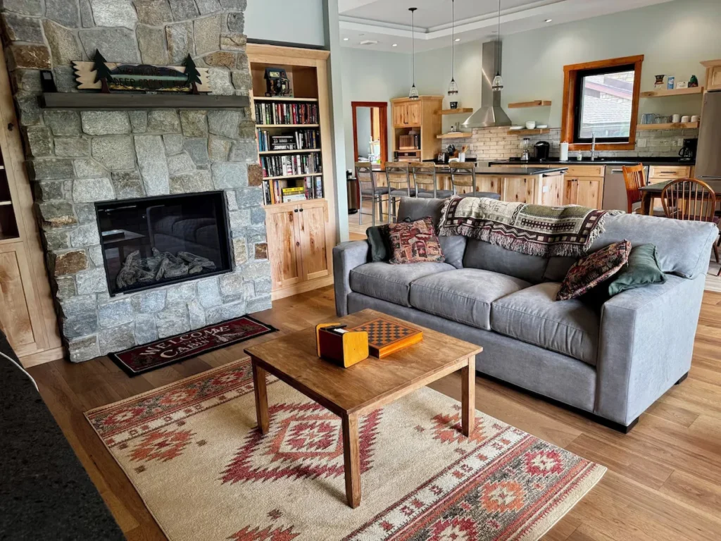 Cabin living room with a large stone fireplace, gray sofa, southwestern rug, and open kitchen in the background