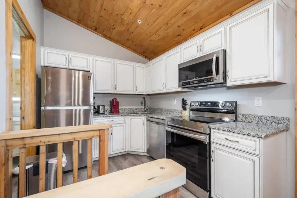Cabin kitchen with white cabinets, granite countertops, and stainless steel appliances under a wood ceiling