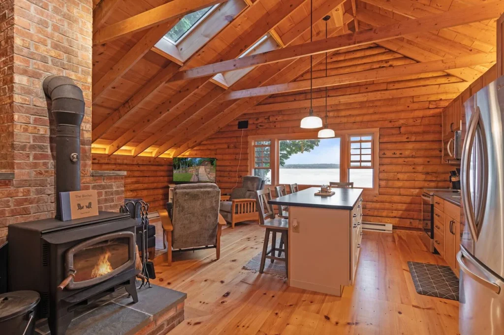 Log cabin interior with a wood stove, kitchen island, vaulted ceilings, and lake views through the window