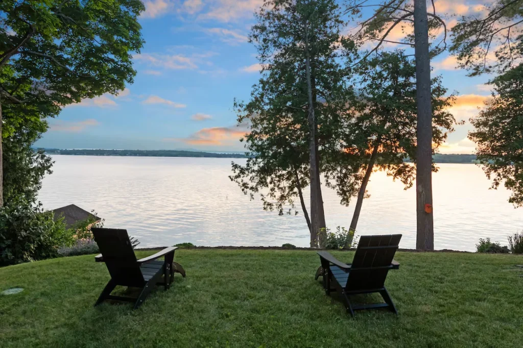 Two black Adirondack chairs on a grassy lawn overlooking a wide lake at sunset
