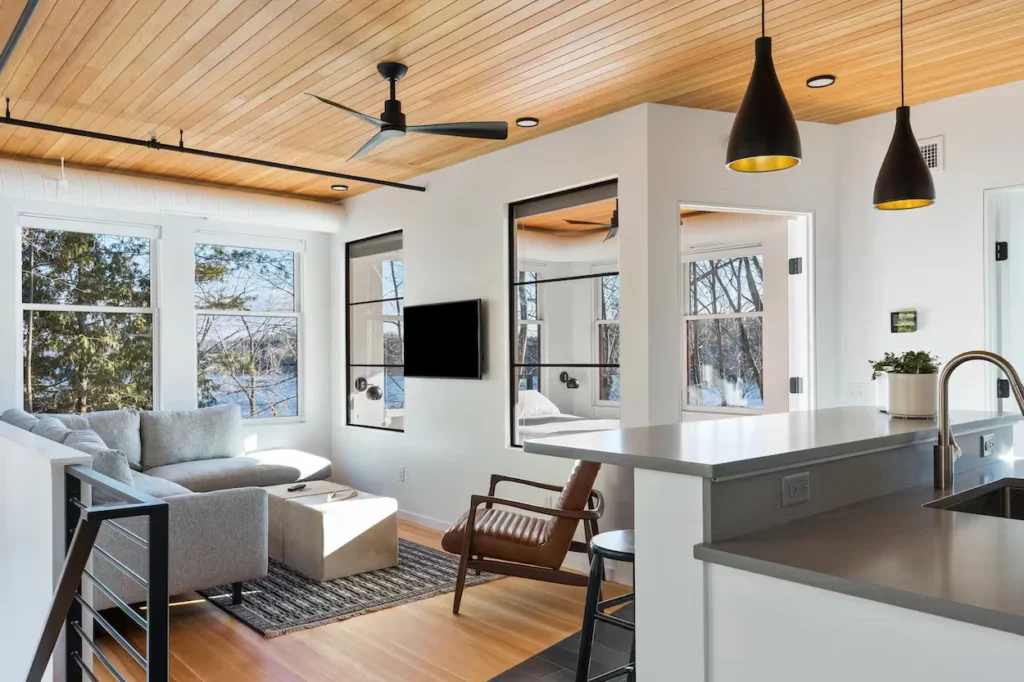 Modern open layout cottage interior featuring a grey sectional sofa, wood paneled ceilings, and kitchen barstools