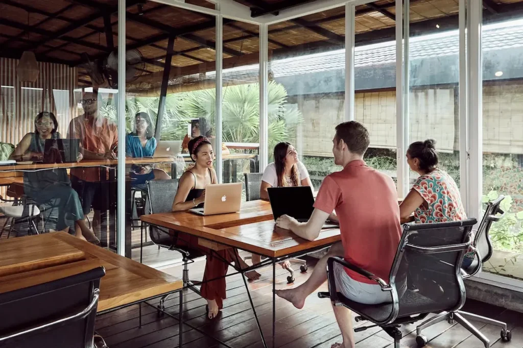 Group of professionals working on laptops and talking around a wooden table inside an Outsite glass-walled coworking office in 2026