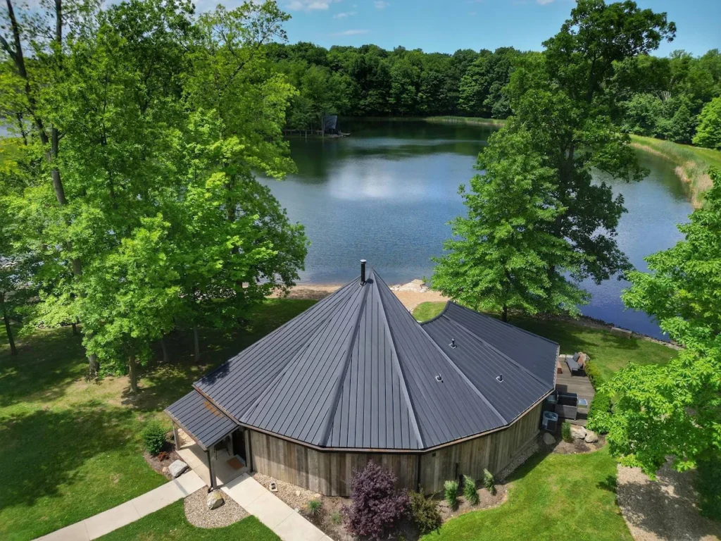 Aerial view of a unique circular cabin with a dark metal roof positioned near a scenic lake surrounded by dense green woods