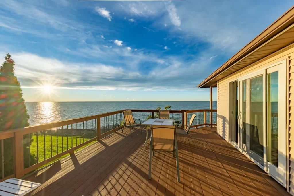 A wooden elevated deck with patio furniture overlooking a wide blue lake during sunrise