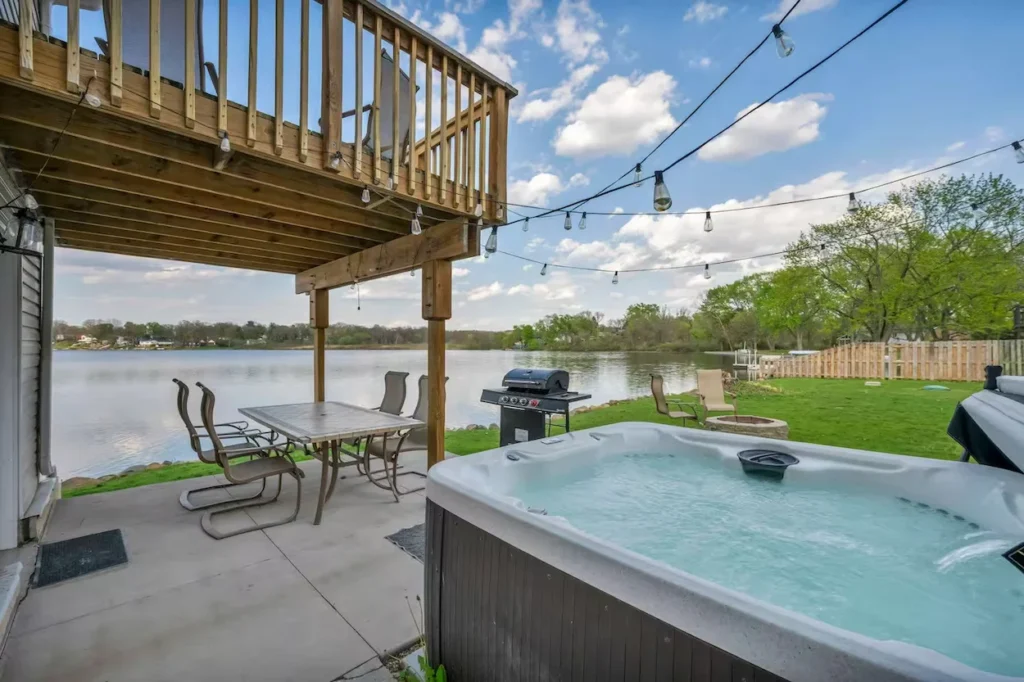 Lakeside concrete patio with a bubbling hot tub, dining set under a wooden deck, and a grill