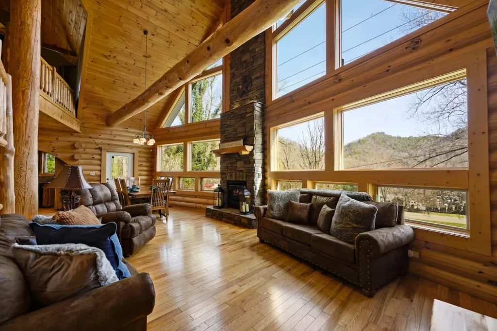 Spacious cabin living room with a stone fireplace, log beams, and large scenic windows