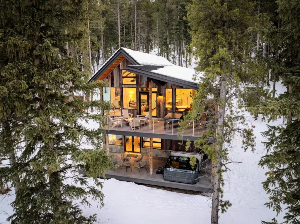 Snow-covered multi-level modern wood cabin in a pine forest with an illuminated interior and deck hot tub