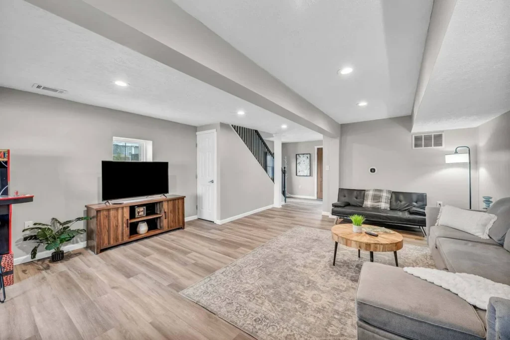 Finished basement living room with gray walls, sectional seating, a wall-adjacent TV stand, and an arcade machine