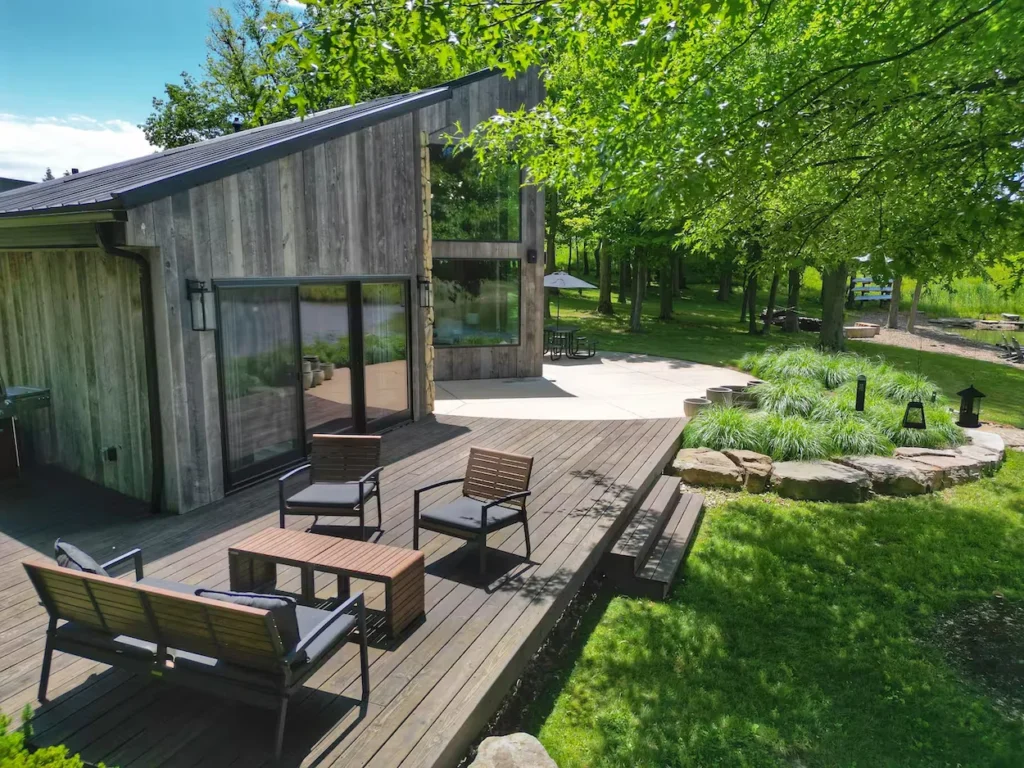 Modern rustic wood cabin with a spacious dark wood deck, outdoor furniture, and a grassy lakeside yard