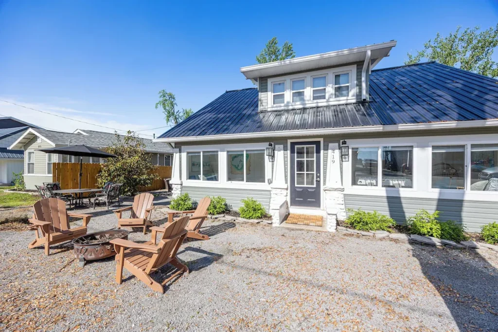 A green lake house cottage with a dark metal roof, sunroom windows, and wooden chairs surrounding a fire pit in the gravel yard