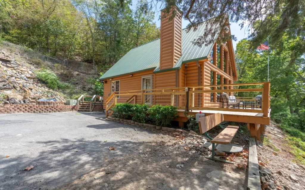 Hot Springs log cabin exterior with a green tin roof, wooden deck, and a parking area flanked by trees