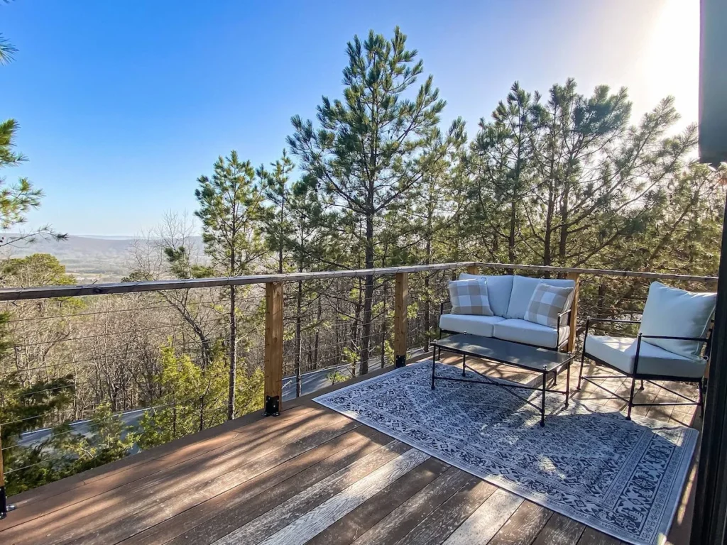 Elevated cabin deck in Hot Springs, Arkansas, with an outdoor rug, loveseat, and wire railings facing dense pine trees