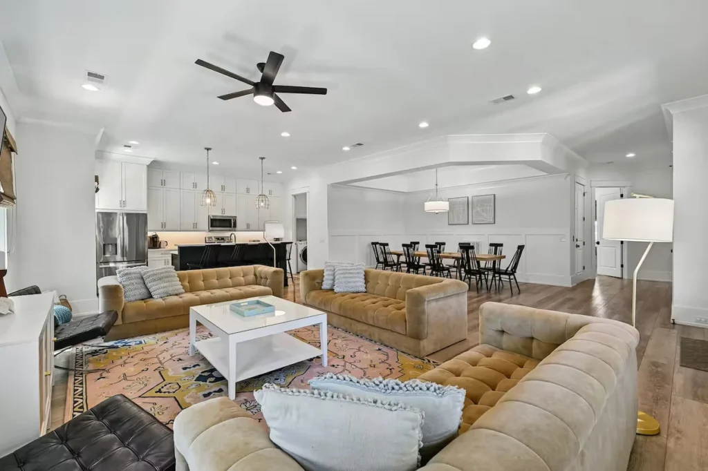 Spacious white living room with tan tufted sofas, a colorful area rug, and an open kitchen and dining area