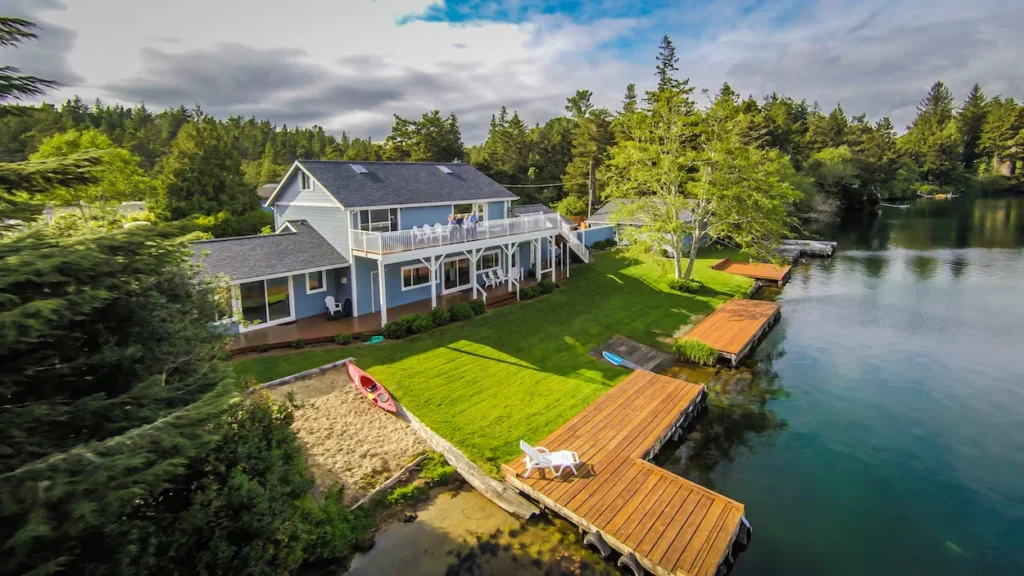 Aerial view of a blue lakefront house with multiple wooden docks, a green lawn, and a red kayak