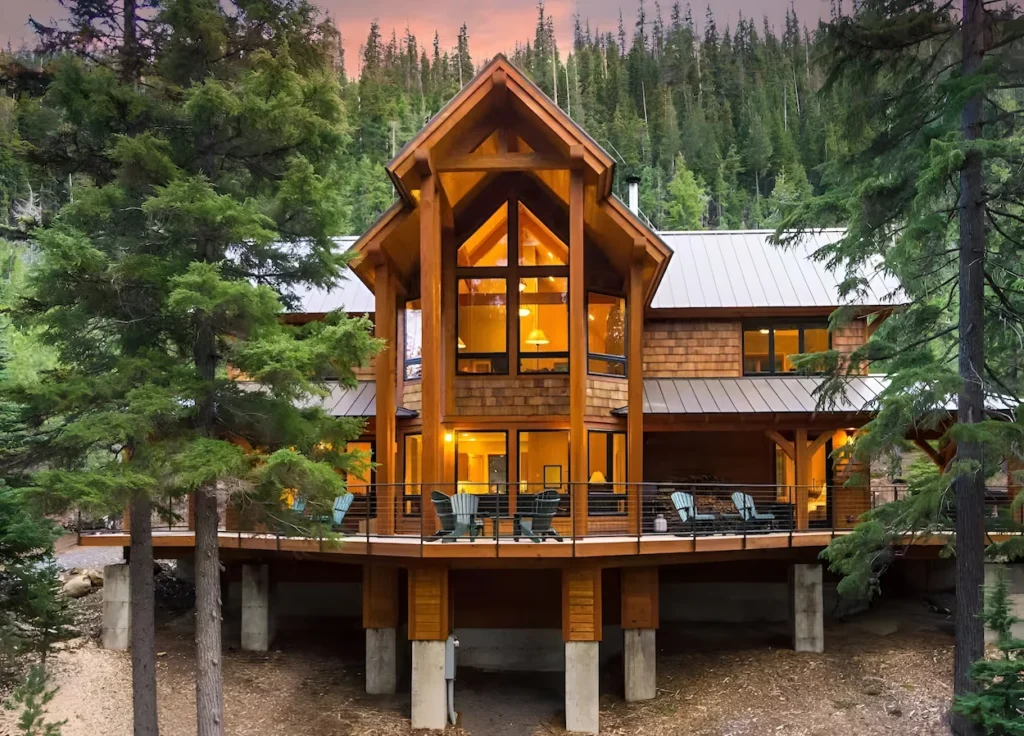 Large timber-frame cabin with glowing windows, a wrap-around deck, and concrete pillars in a pine forest