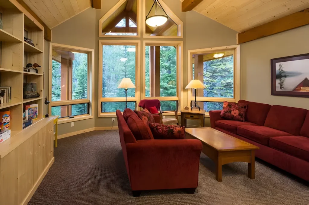 Cabin living room with red sofas, a vaulted ceiling, large windows facing pine trees, and a wooden bookshelf