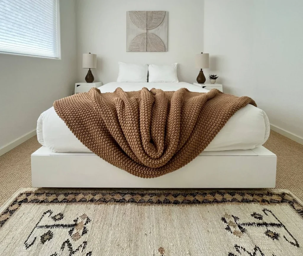 Minimalist white bed with a textured brown knit blanket and a patterned boho area rug
