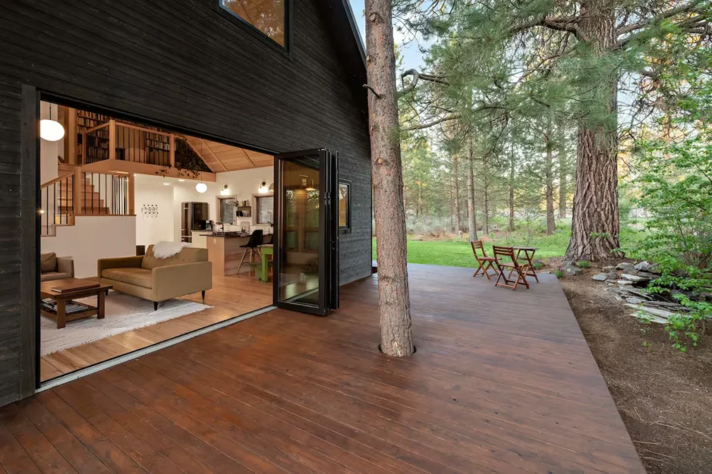 Modern dark wood cabin with open folding glass doors leading to a wooden deck surrounded by pine trees
