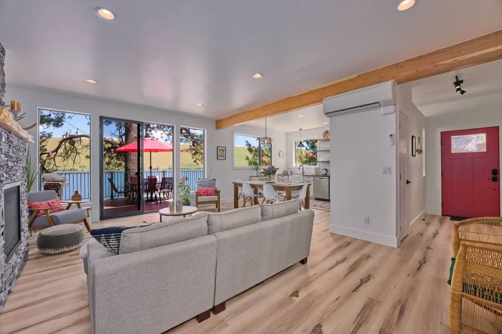 Bright cabin open-concept living room with gray sofa, stone fireplace, and sliding doors to a deck