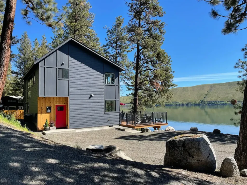Modern dark gray two-story cabin with a red door and a lakefront deck surrounded by pine trees