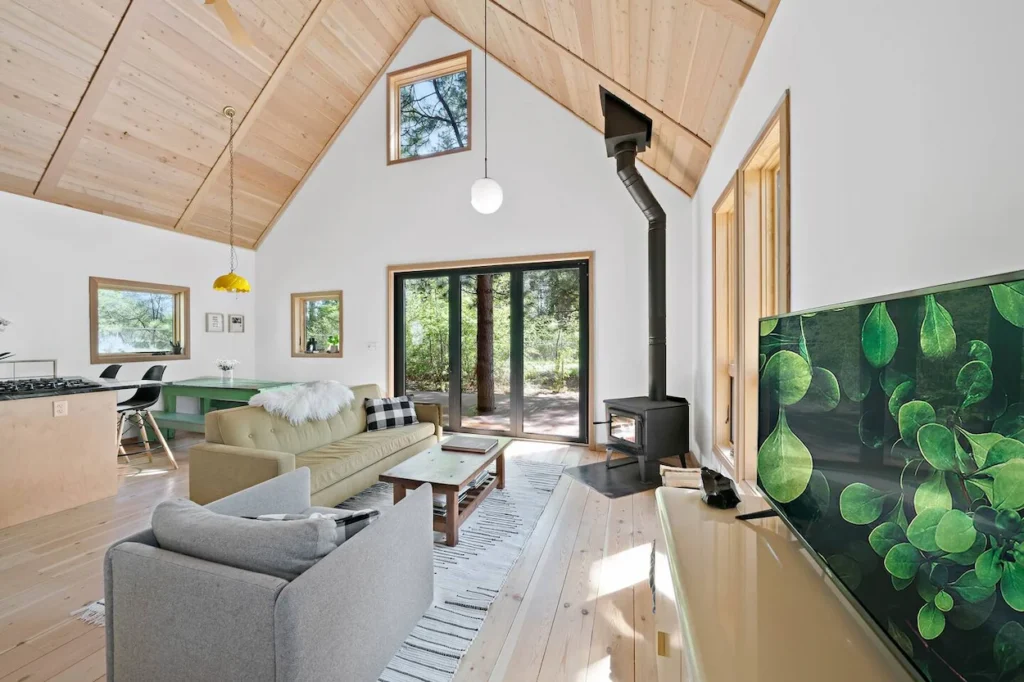 Bright cabin interior with vaulted wood-plank ceilings, a modern wood stove, a beige sofa, and sliding glass doors
