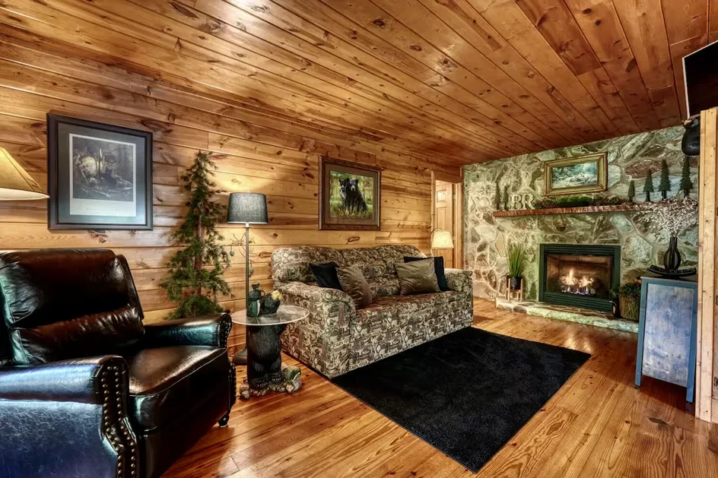 Cabin living room with wood paneled walls, a patterned sofa, leather armchair, and stone fireplace