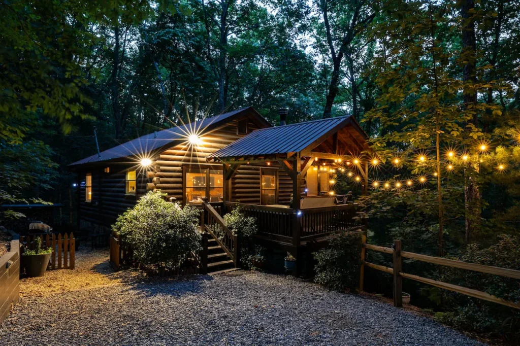 Log cabin with string lights on the porch and a gravel driveway at dusk