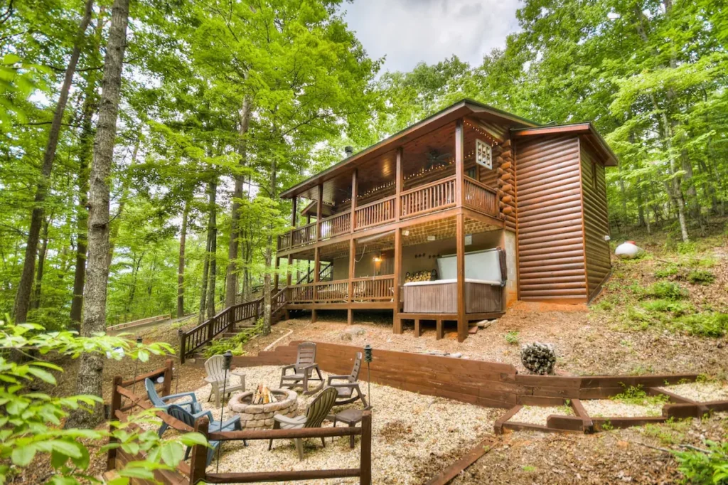 Two-story log cabin with a hot tub on the porch deck and a stone firepit area with chairs