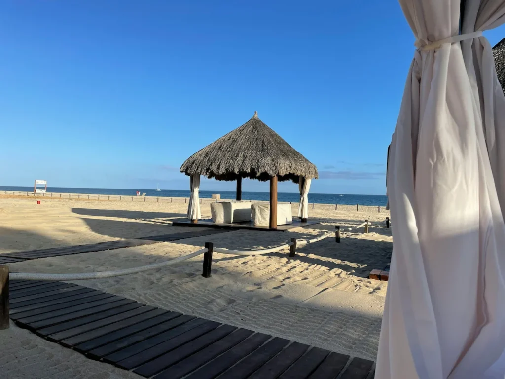A thatched palapa cabana with white curtains on a sandy beach next to a wooden boardwalk