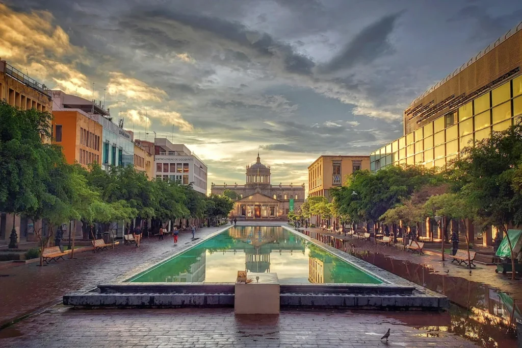 A long rectangular reflecting pool leading toward the historic Hospicio Cabañas building under a cloudy sky