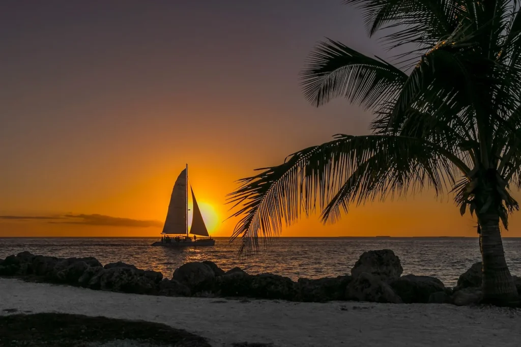 A sailboat silhouetted against a bright orange sunset behind a palm tree in Key West, Florida