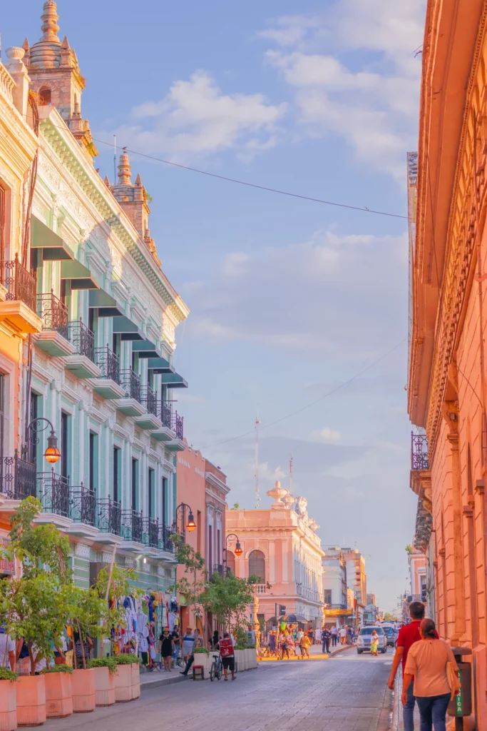 Pedestrians walking down a sunny street lined with pastel-colored colonial buildings in Mérida, Mexico