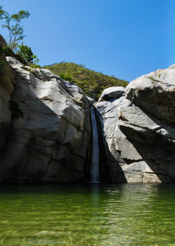 A tall waterfall cascading down massive gray rocks into a clear green swimming pool