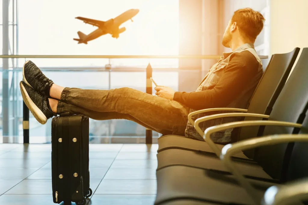 Man at an airport terminal with feet on a carry-on suitcase, holding a phone and watching a departing airplane