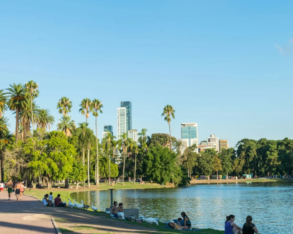 People and white geese sit on a grassy bank along a lake lined with palm trees and a city skyline backdrop