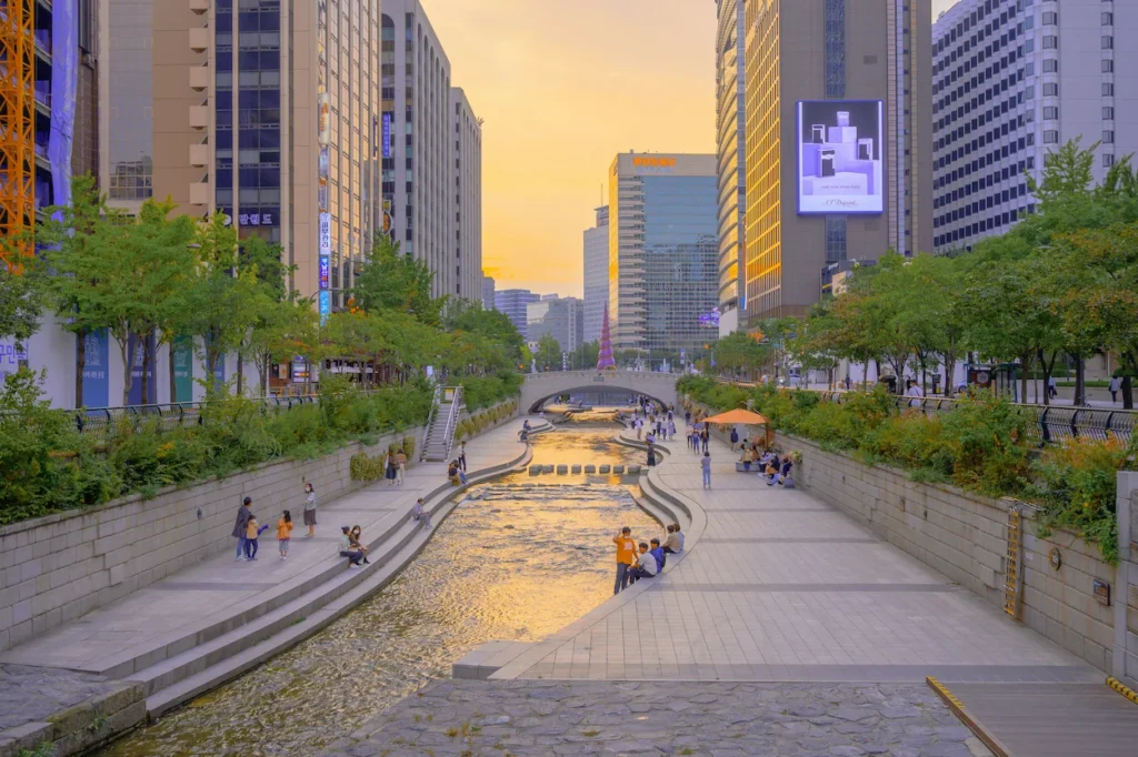 Cheonggyecheon Stream lined with walking paths and lush greenery under an urban bridge at sunset