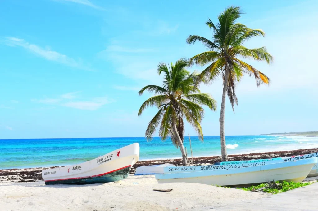 Two fishing boats parked on a sandy beach next to turquoise water and palm trees in Cozumel
