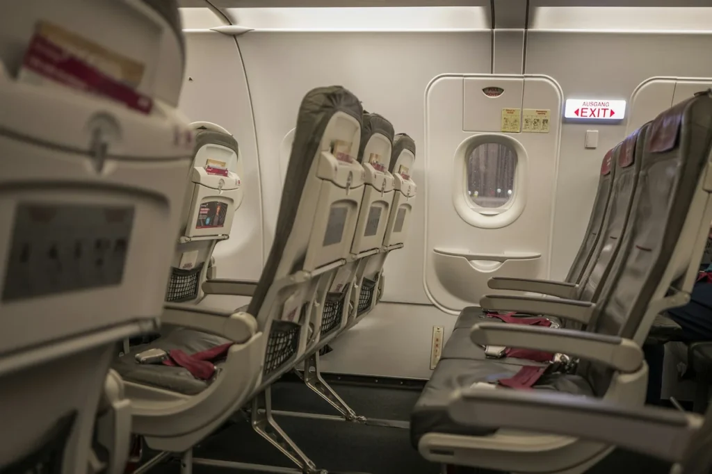 Empty grey slimline airplane seats in an exit row next to a window and a German "Ausgang Exit" sign