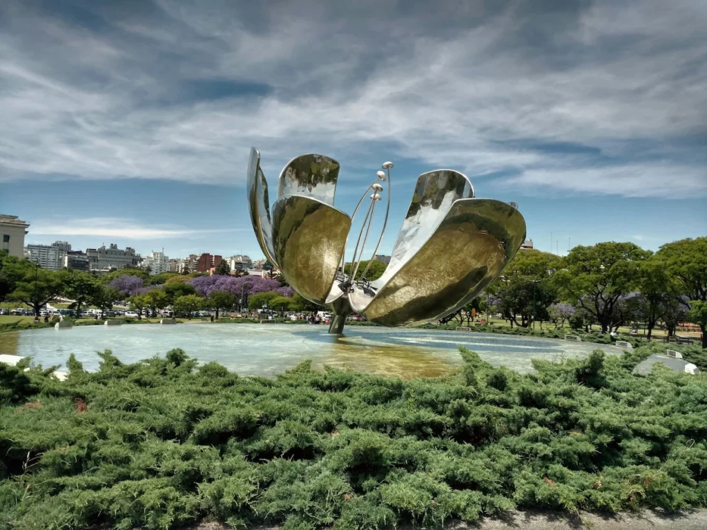 Large metallic flower sculpture opening over a reflection pool surrounded by green trees in Buenos Aires