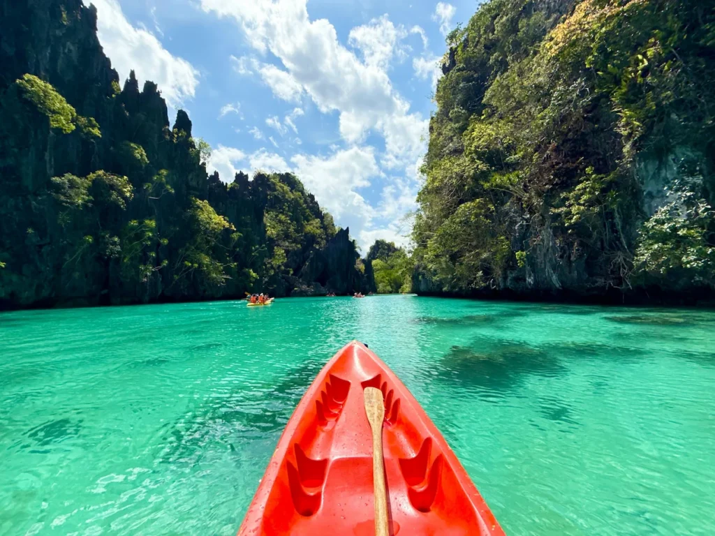 First-person view from a red kayak paddling through a clear turquoise lagoon with towering limestone cliffs