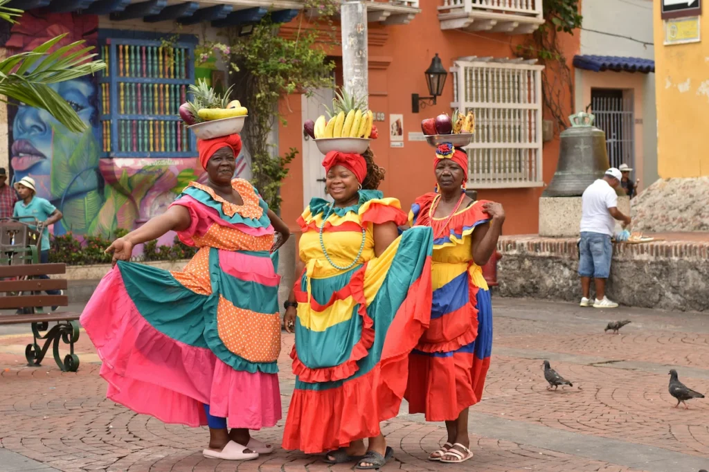 Three Palenquera women in colorful dresses balancing bowls of fresh fruit on their heads