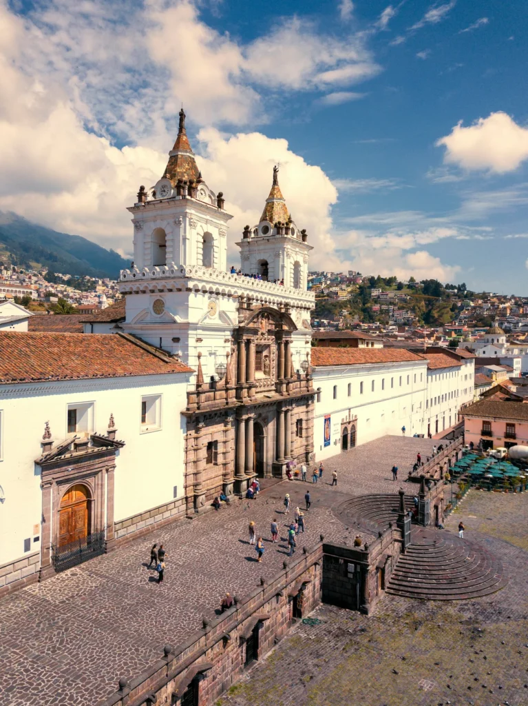 White twin towers of San Francisco Church overlooking a stone plaza with pedestrians in Quito