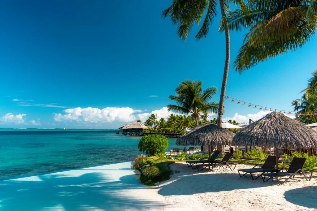 Lounge chairs under thatched umbrellas on a white sand beach next to a turquoise ocean and an infinity pool