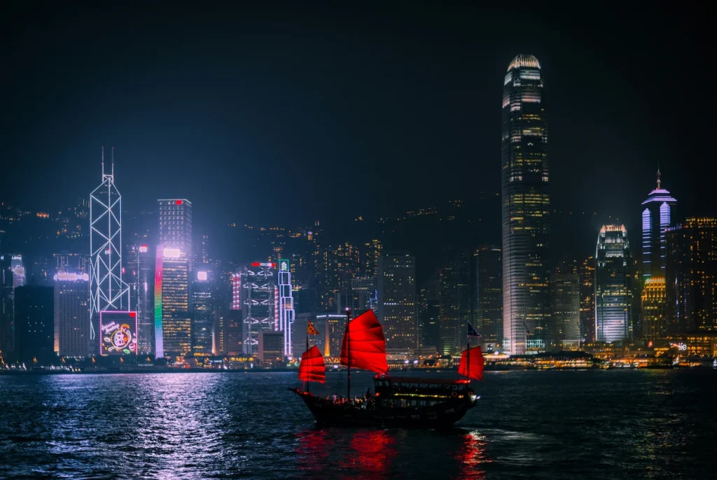 Traditional red-sailed junk boat sailing across Victoria Harbour at night with illuminated Hong Kong skyscrapers