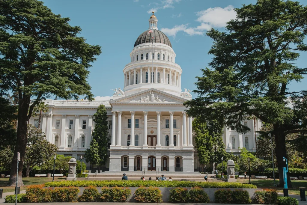 The neoclassical white dome and facade of the California State Capitol building framed by green trees